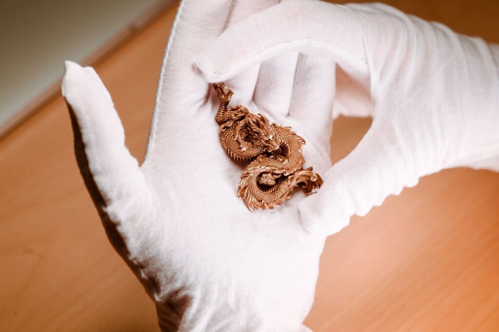 Close-up of a handcrafted dragon pendant held in white gloves, showcasing intricate metalwork.